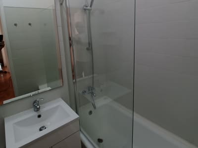 Casa de banho remodelada, com resguardo de vidro sobre a banheira, móvel de lavatório novo e revestimento branco.